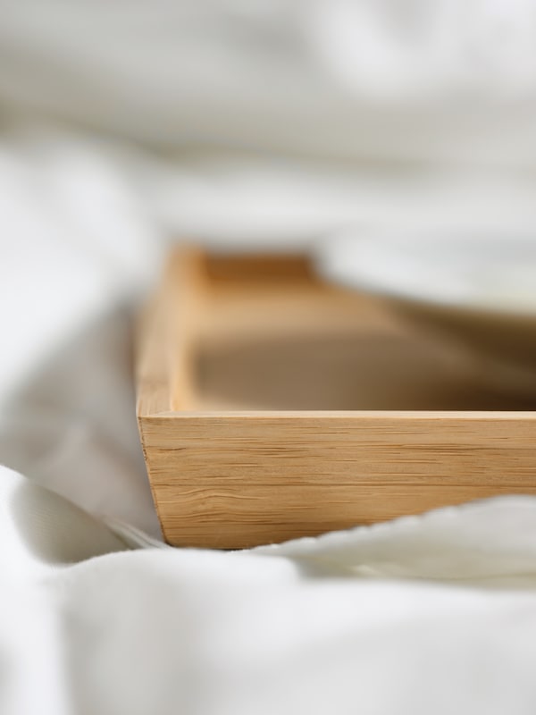 One corner of an OSTBIT bamboo tray on white bed linen, showing the raised edge, and with a white plate on it.