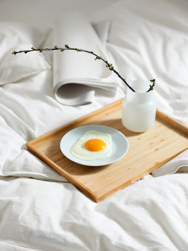 An OSTBIT tray on top of white bed linen, holding a white vase with a twig in it, and a small plate with a fried egg on it.