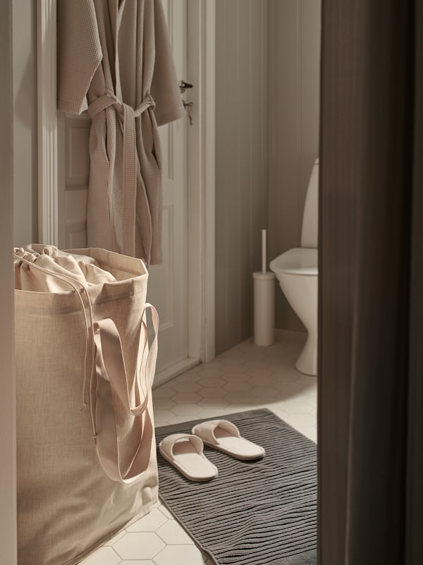 A beige PURRPINGLA laundry bag and a pair of slippers are near a bathroom door with a beige BJÄLVEN bath robe hanging on it.