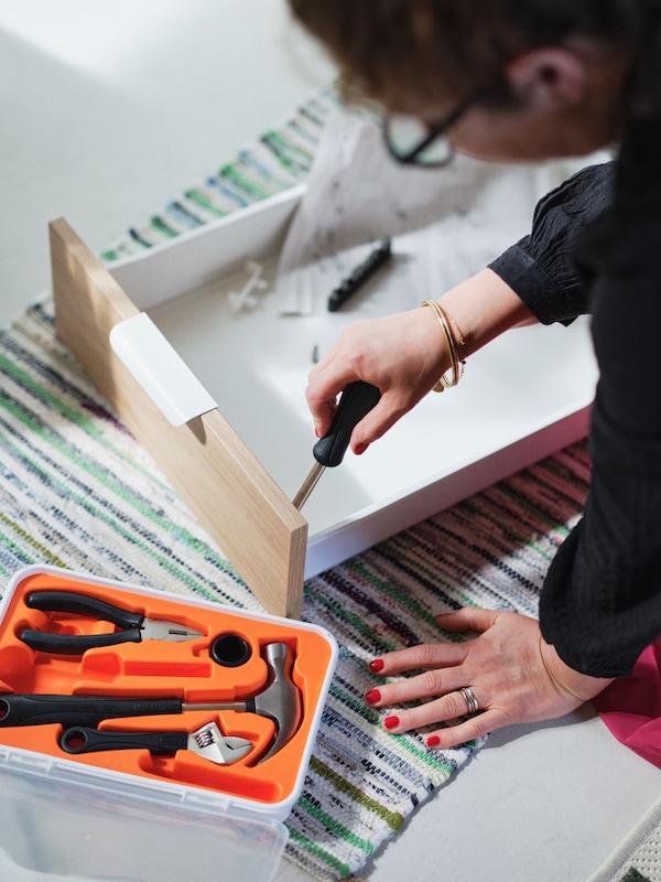 A woman is sitting on the floor while putting together a drawer using a screwdriver from the FIXA 17-piece tool set.
