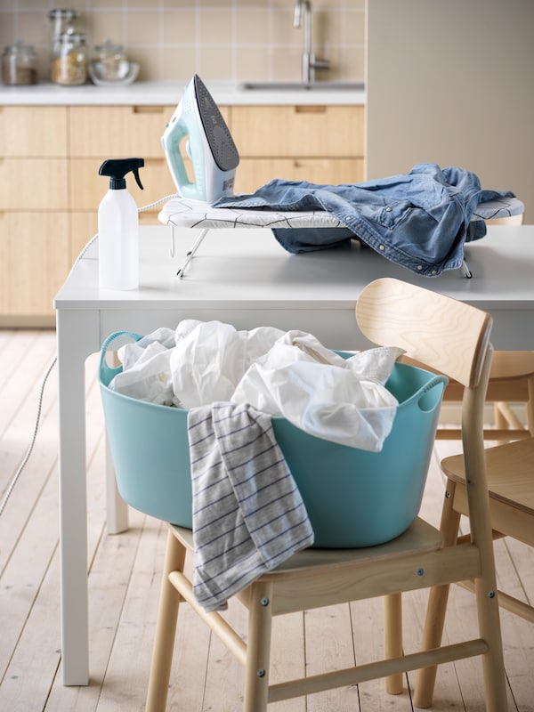 A light-blue TORKIS laundry basket, a JÄLL table ironing board and other laundry-care items set up around a kitchen table.