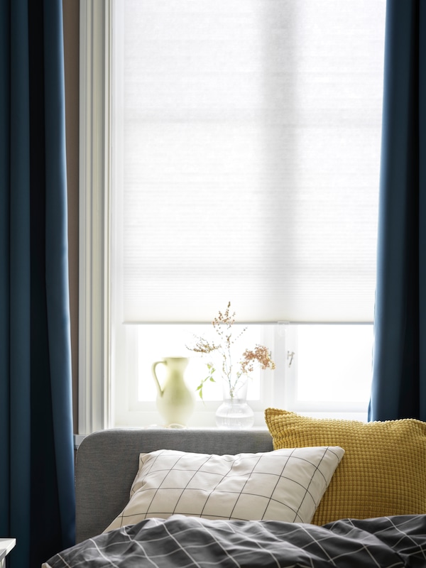 The head of a bed beside a window with a window treatment consisting of a HOPPVALS cellular blind and HILLEBORG curtains.