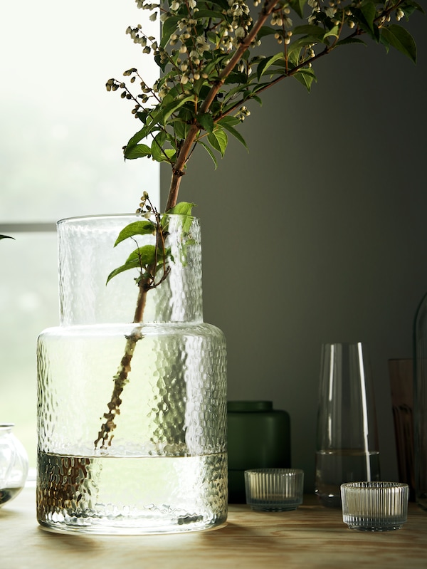 A KONSTFULL vase made in clear glass with a bubbly pattern, holding a decorative branch of greenery by a window.