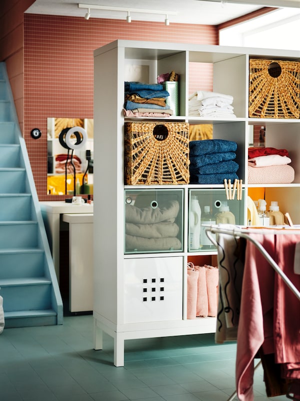 A white KALLAX shelving unit with underframe used as a room divider and storage for laundry items and textiles.