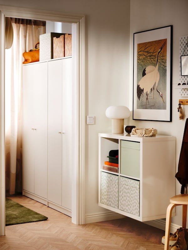 A KALLAX shelving unit with DRÖNA boxes is mounted to the wall in a hallway near a bedroom with two KLEPPSTAD wardrobes.