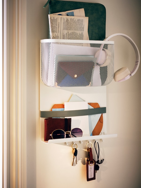 A white DRÖNJÖNS wall storage with hooks hangs on a wall with keys, documents, and accessories in it.
