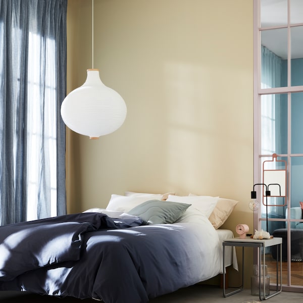 A RISBYN pendant lamp hangs over a bed with STRANDTRIFT bed linen next to a window with HILJA curtains.
