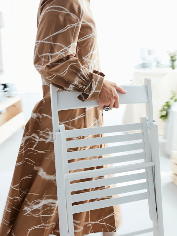 A woman with a brown patterned dress carrying a TERJE folding chair in white wood in a white setting.