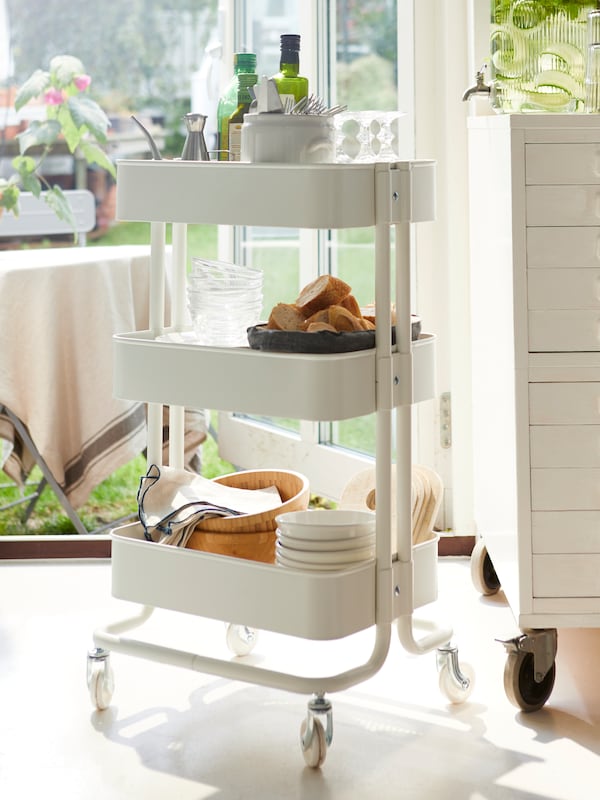 The three shelves of a RÅSKOG kitchen cart are full with a bowl of bread, dishes, napkins and more.