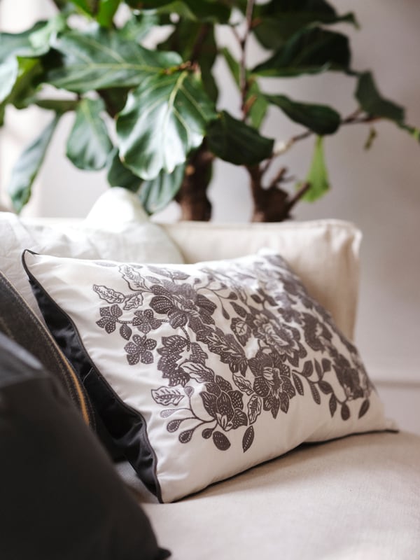 A SKUGGNÄVA white and grey cushion with dark grey florals sits on a sofa, with a large plant in the background.