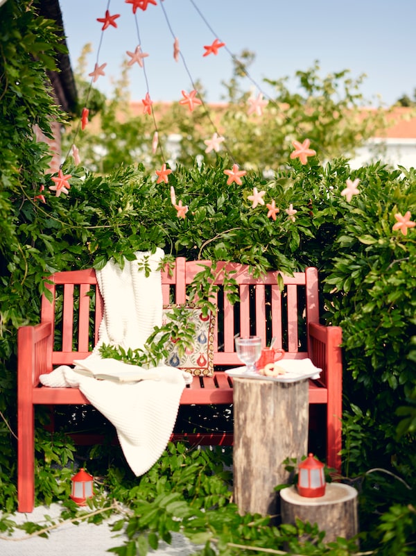 A red PÄRONHOLMEN bench with backrest stands in a garden with SOLVINDEN LED lighting chains hanging above.