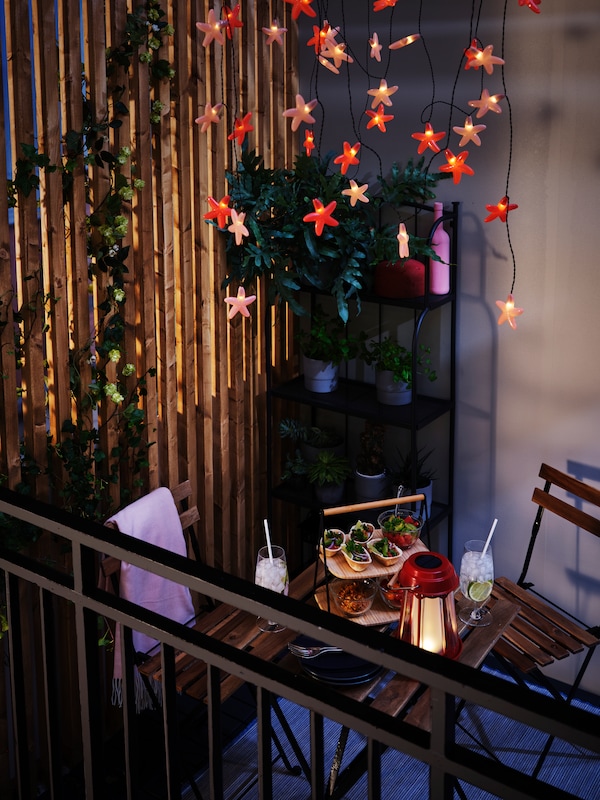 Food, drinks and a SOLVINDEN LED table lamp are on a TÄRNÖ table on a balcony with SOLVINDEN lighting chains hanging above.