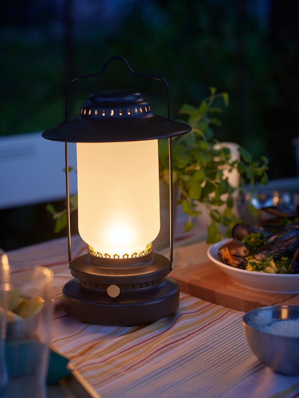On a summer night, a STORHAGA LED table lamp illuminates a table set for dinner with SOLMOTT fabric used as a tablecloth.