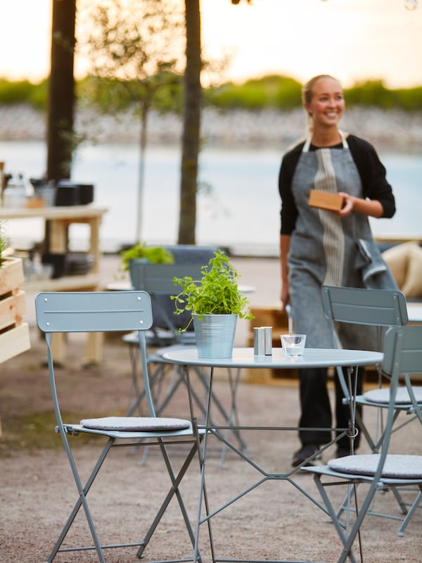 A smiling waitress carrying a small box at an outdoor café with grey tables and chairs.
