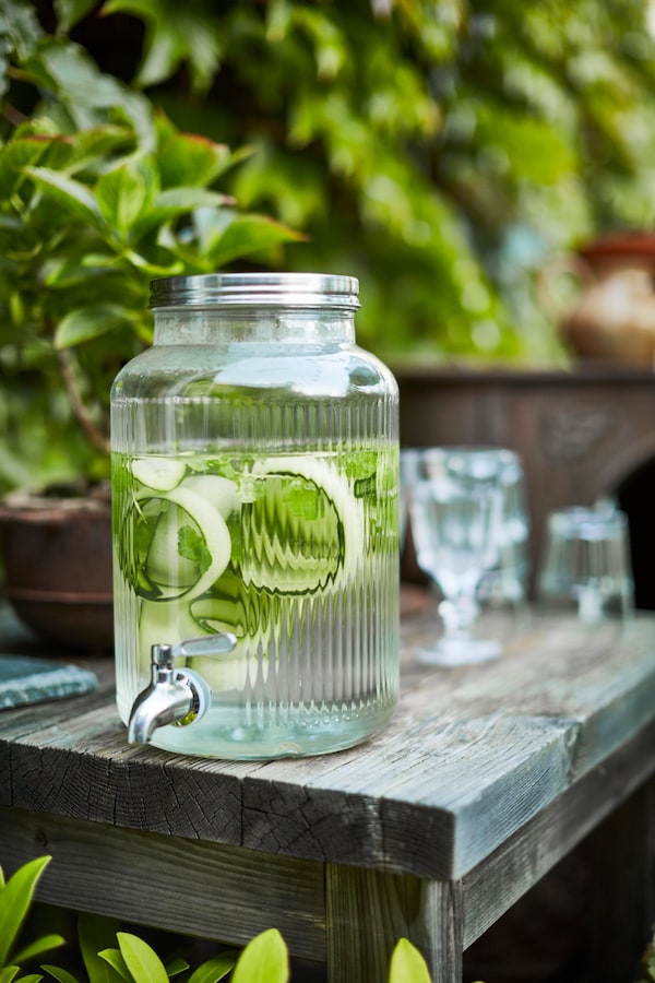A VARDAGEN jar with tap containing water and strips of cucumber stands at the edge of a table on a terrace.