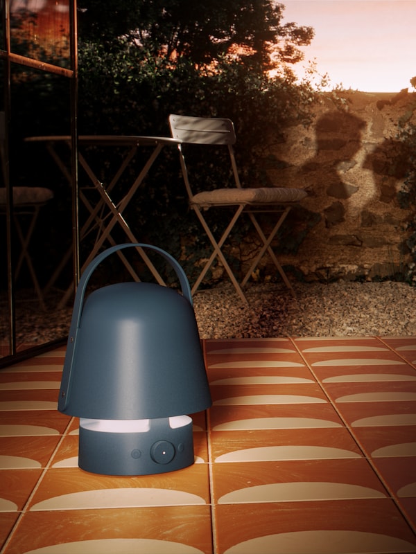 A VAPPEBY Bluetooth speaker lamp stands on a tiled part of an outdoor area near a wall with the shadow of a couple on it.