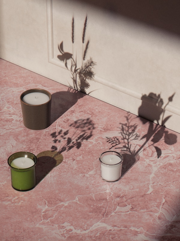 ADLAD, HEDERSAM and ENSTAKA scented candles in jars stand on a marbled surface with flower-shaped shadows.