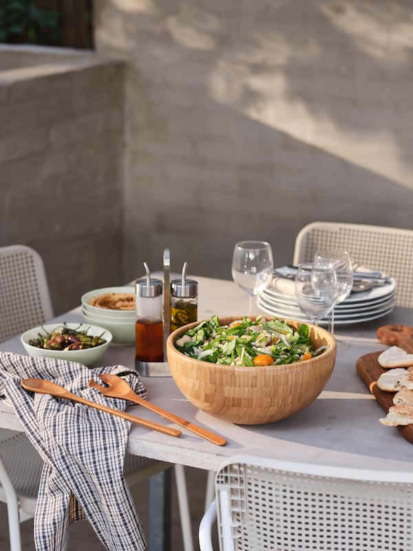 An outdoor, sunlit table set with salad in a BLANDA MATT bamboo serving bowl, SVALKA wine glasses and other tableware.