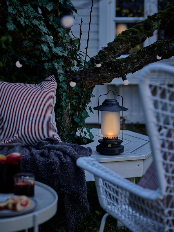 A lit STORHAGA dimmable LED table lamp placed on a bench next to a white HÖGSTEN armchair on a patio in dim light.