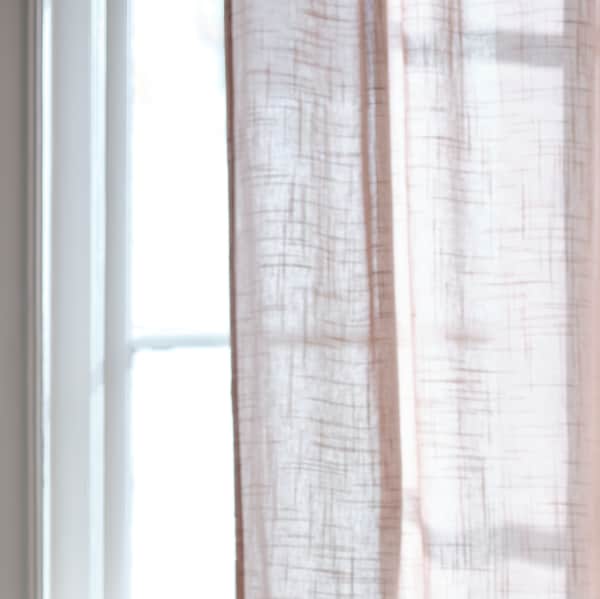 Part of a light pink SILVERLÖNN sheer curtain which is hanging in a bright window on a sunny day.