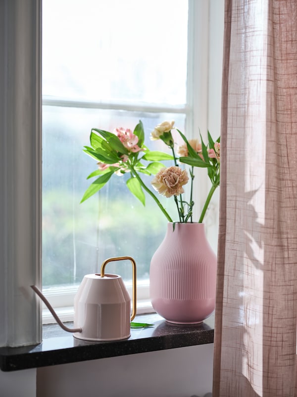 A pink GRADVIS vase with flowers and a pale-pink VATTENKRASSE watering can on the sill of a window with sheer curtains.