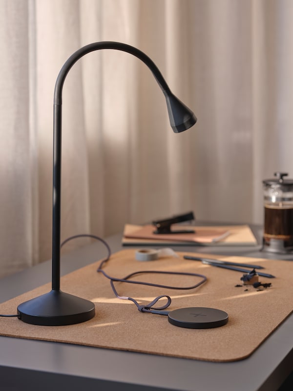 A black NÄVLINGE LED work lamp stands on a cork SUSIG desk pad on a desk near a black LIVBOJ wireless charger.