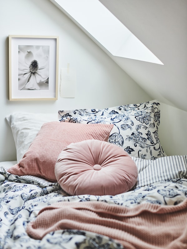 A bed in a room with a sloping ceiling with JUNIMAGNOLIA white/dark blue bed linen and pink cushions and a throw.