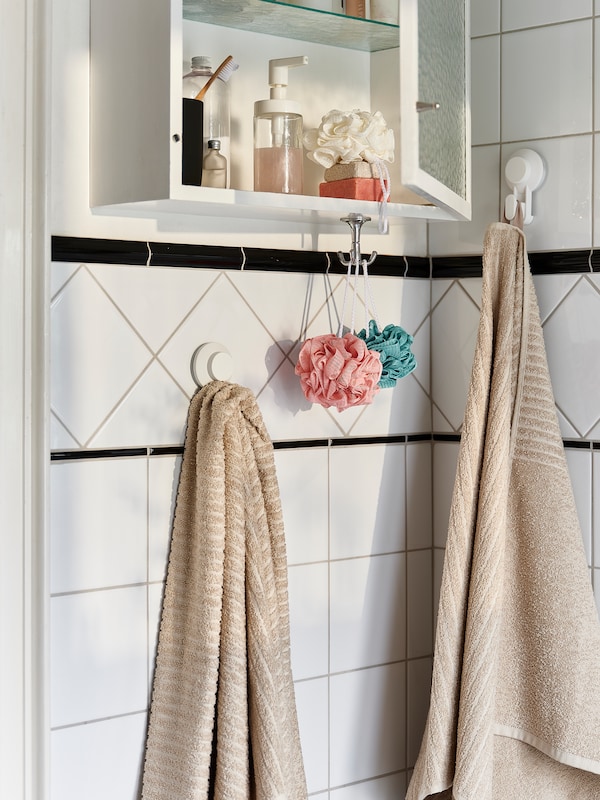 Light-beige VÅGSJÖN bath towels hanging from TISKEN suction-cup hooks in a white-tiled bathroom.