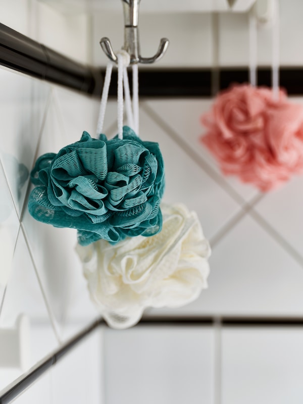 A three-piece set of ÅBYÅN multicolour body puffs hanging on hooks in a white-tiled bathroom.