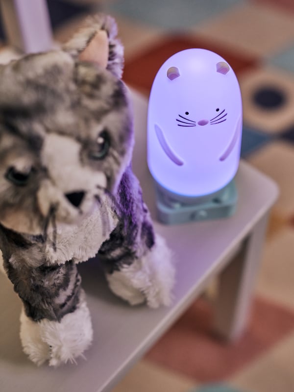 A chair with a lit SPIKEN otter-shaped LED night light placed next to a grey and white LILLEPLUTT soft toy cat.