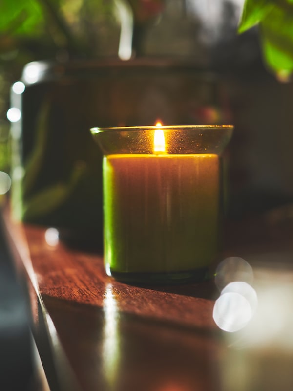 A lit HEDERSAM fresh-grass scented candle placed on a dark-wood surface in a dimly lit room.