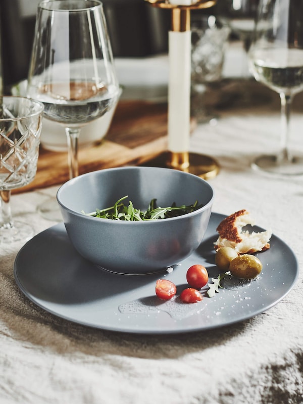 Plateware holding greens and fruit on a rustic table mat next to elegant wine glasses with white wine. 