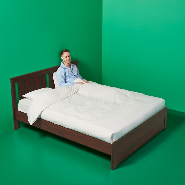The corner of a green room with a dark wooden bed in it covered in white bed linen and with a man sitting up in it.