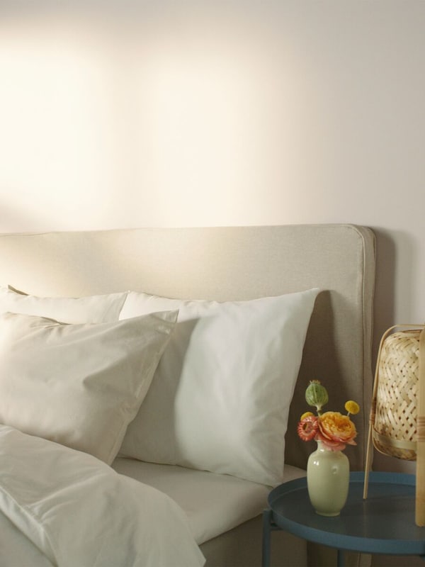 The top of a bed with headboard, dressed with bedding and pillows with a blue side table with a lamp and flowers.