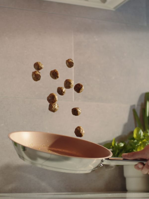 Twelve, HUVUDROLL plant balls suspended in mid-air above a shiny, stainless-steel, frying pan in front of a beige wall.