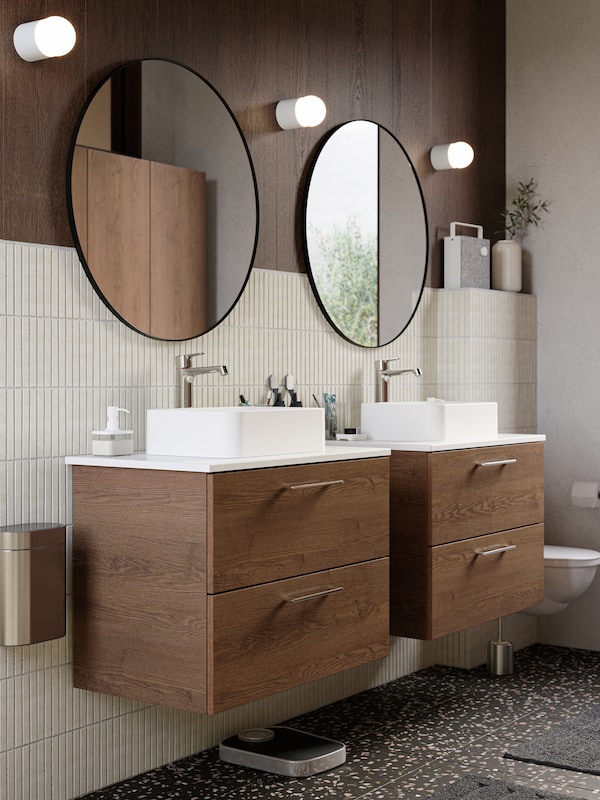 Two brown stained ash effect wash-stands, two large round mirrors, three wall lamps, beige tiles and dark terrazzo flooring.