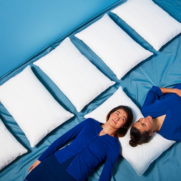 Two people wearing dark blue clothes lie on a blue floor with their heads on a pillow, beside a row of other pillows.