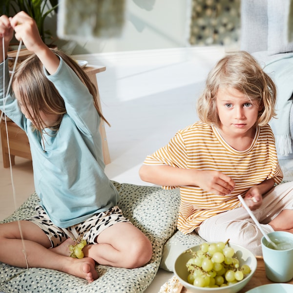 Two young children sitting on the floor in a living room in light-green tones making grape necklaces.