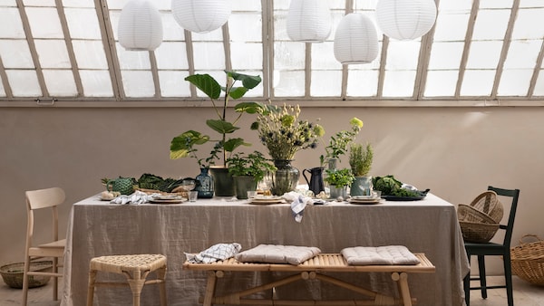Under a glass roof is a table with a linen tablecloth, plates, and plenty of plants and branches in plant pots and vases.
